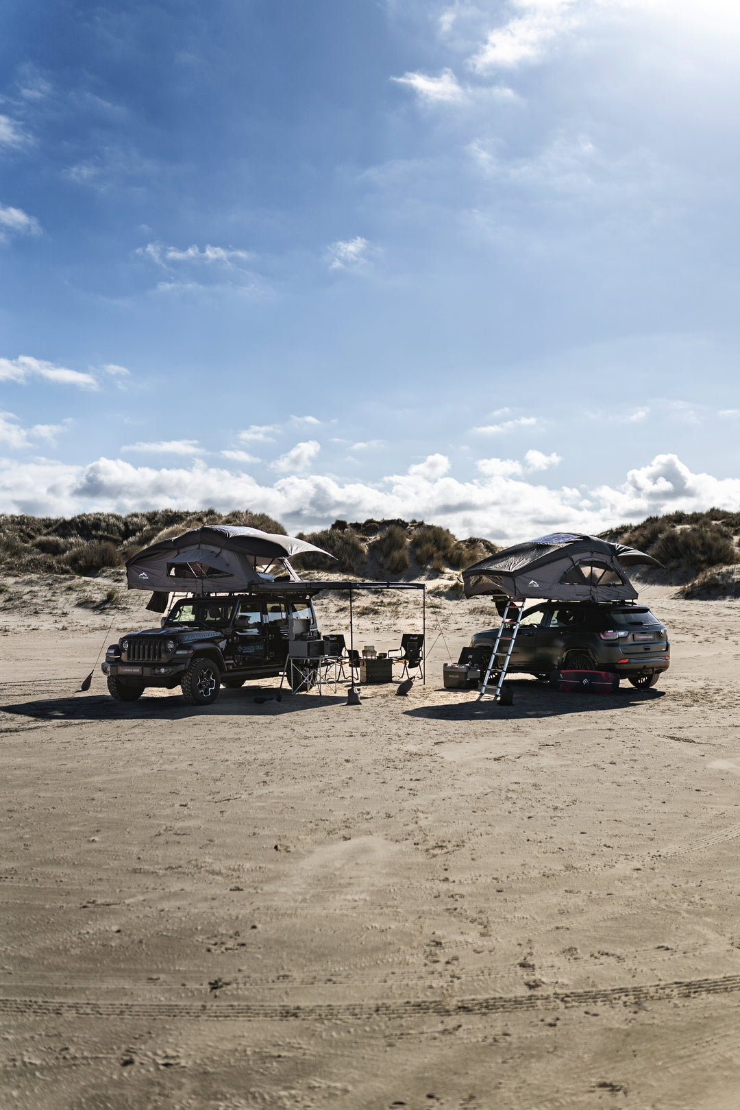 Zwei Geländewagen mit aufgebauten Dachzelten und Outdoor-Setup am Strand unter blauem Himmel.